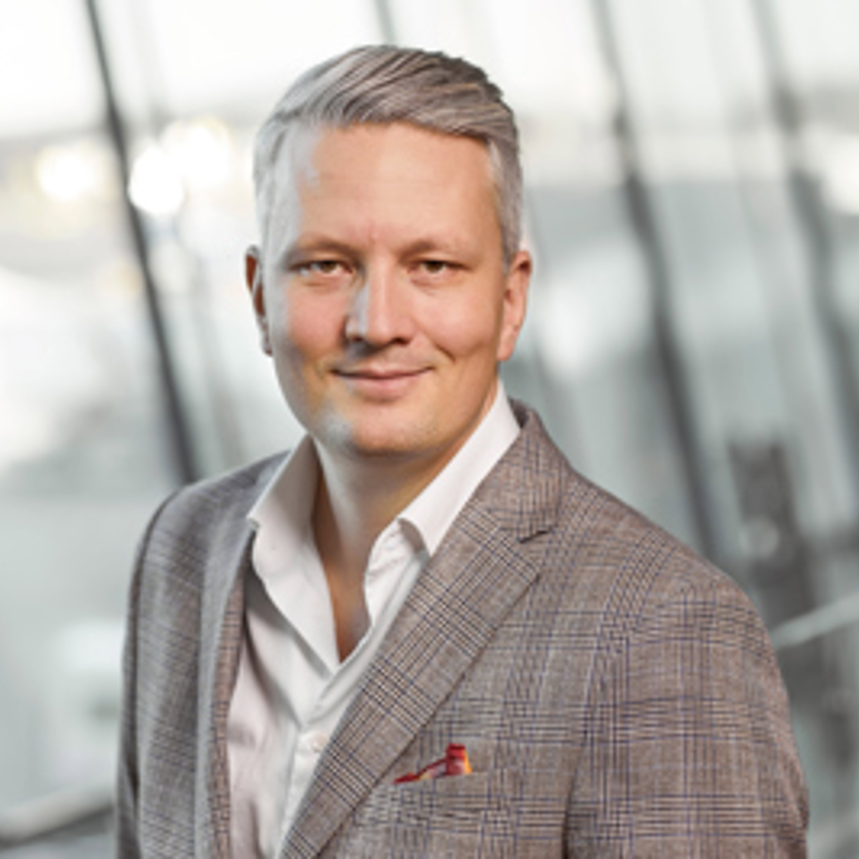 Smilende mann i en grå, rutete blazer som står i en moderne bygning med glassvegger