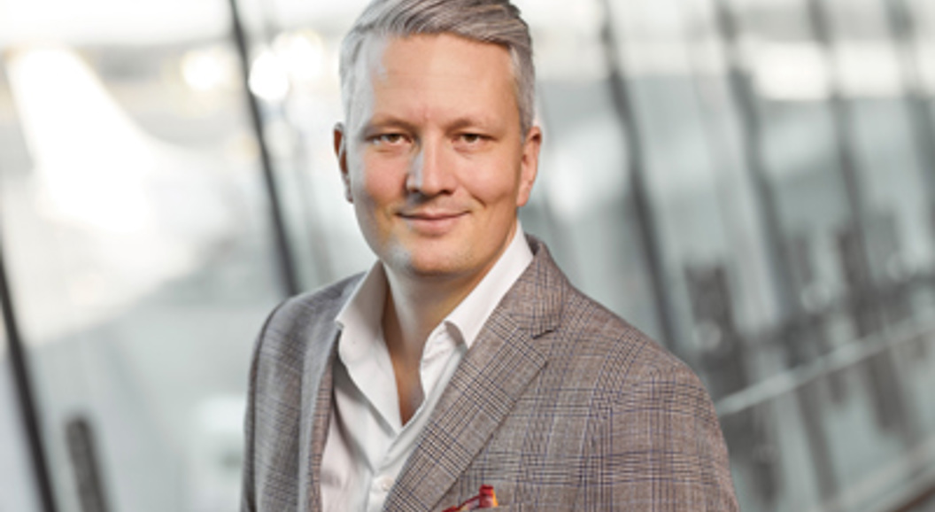 Smilende mann i en grå, rutete blazer som står i en moderne bygning med glassvegger