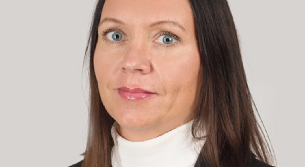 Profesjonell kvinne med langt mørkt hår iført en hvit høyhalset genser og svart blazer, ser direkte på kameraet mot en ensfarget grå bakgrunn