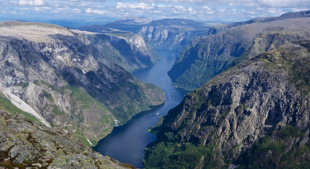 Majestetisk utsikt over en dyp blå fjord omgitt av ruvende steinete klipper og frodige grønne landskap under en klar blå himmel med spredte skyer i Norge