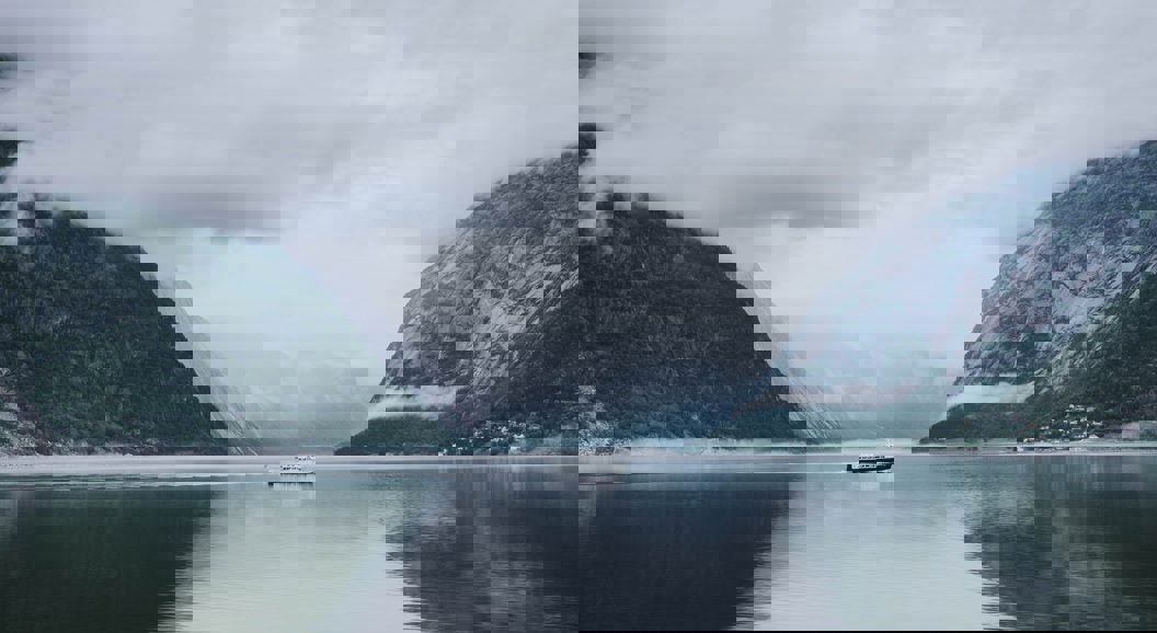 Et rolig landskap med en båt på en stille fjord omgitt av tåkete fjell, under en skyet himmel