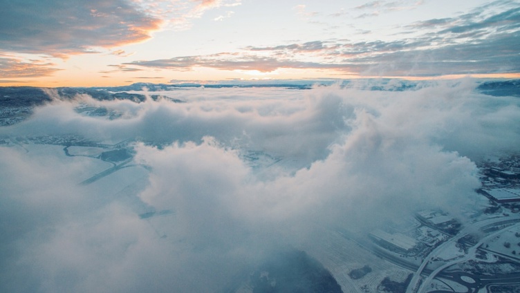 Luftfoto av et snødekt landskap med tett skydekke og en oransje-rosa soloppgang i horisonten