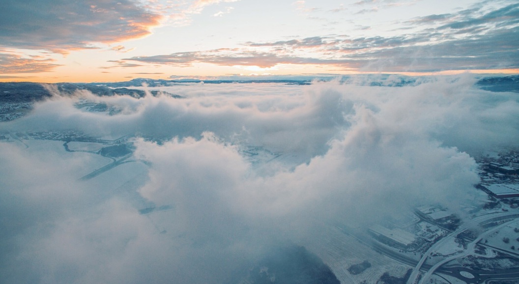 Luftfoto av et snødekt landskap med tett skydekke og en oransje-rosa soloppgang i horisonten