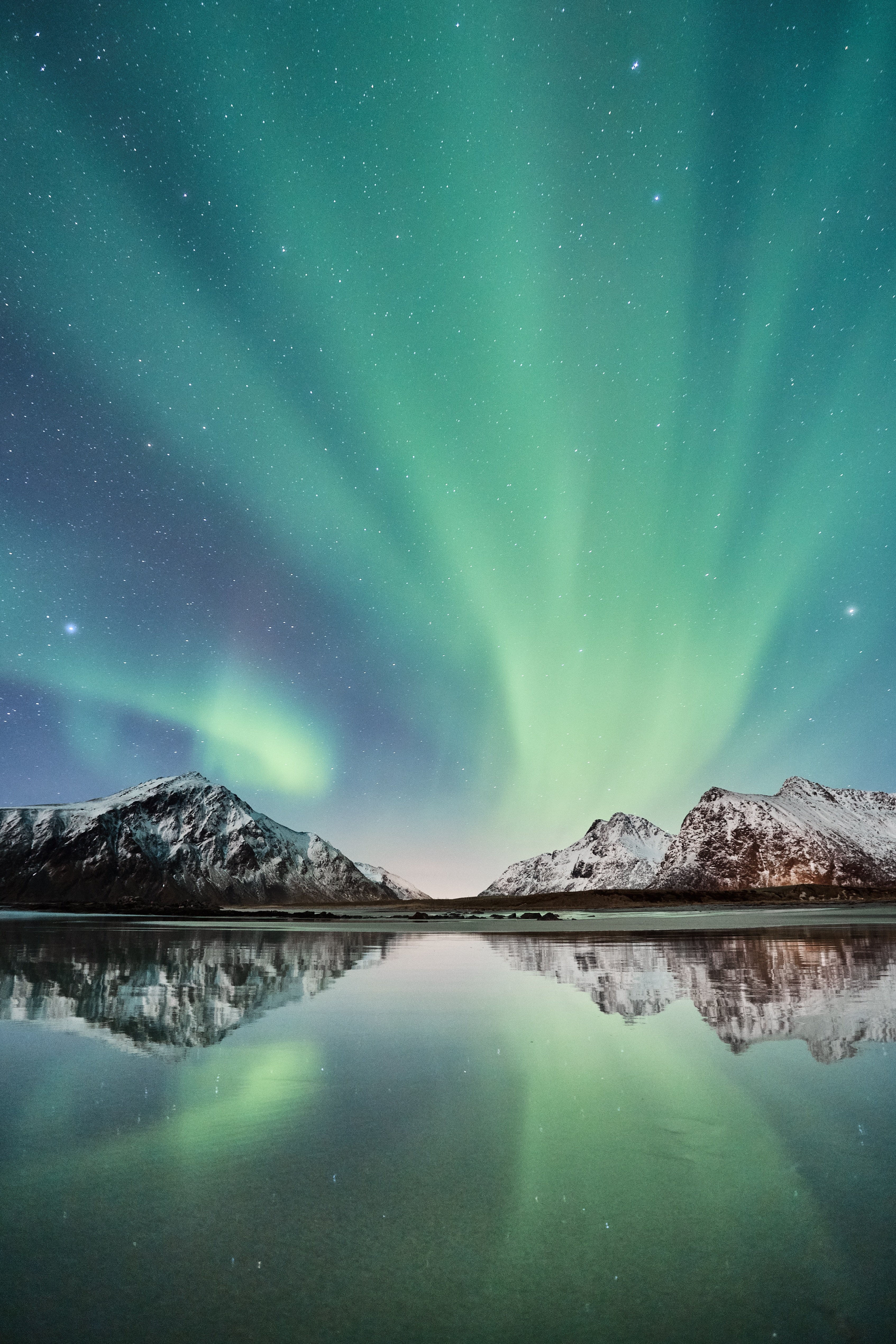 Nordlys over snødekte fjell ved en rolig innsjø i Norge. Refleksjonene av nordlyset i vannet skaper et spektakulært lysbilde.