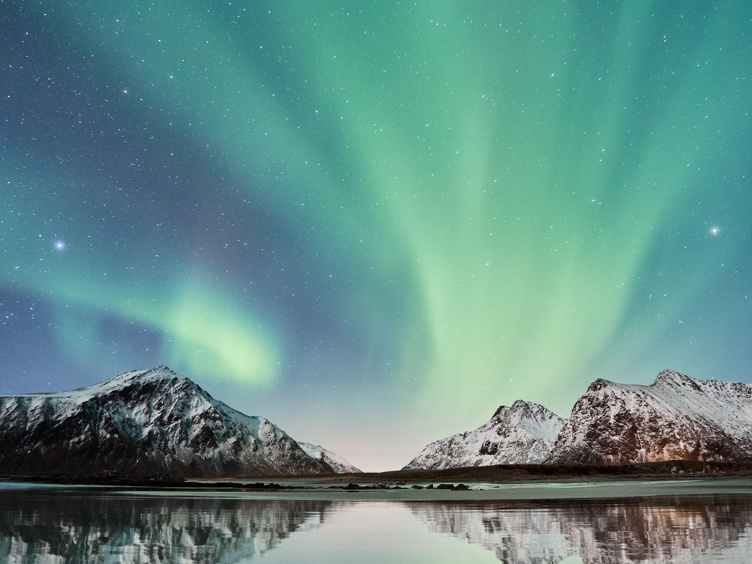Nordlys over snødekte fjell ved en rolig innsjø i Norge. Refleksjonene av nordlyset i vannet skaper et spektakulært lysbilde.