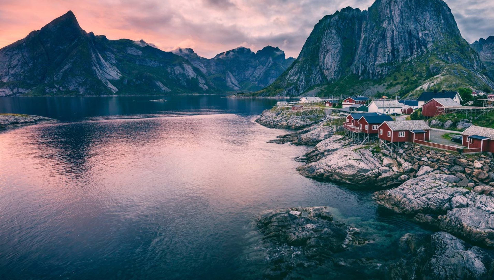 Fjordlandskap i Lofoten med røde rorbuer ved havet og majestetiske fjell i bakgrunnen under en rosa solnedgang.