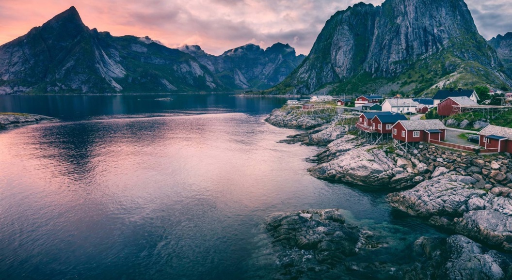 Fjordlandskap i Lofoten med røde rorbuer ved havet og majestetiske fjell i bakgrunnen under en rosa solnedgang.