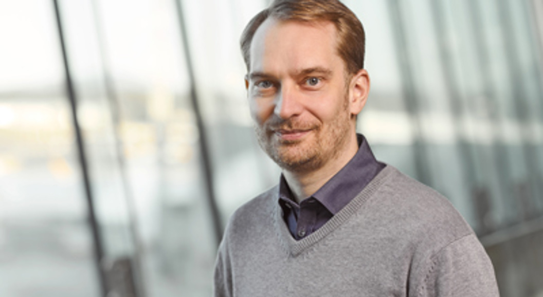 En smilende mann med skjorte og genser står foran en glassvegg.