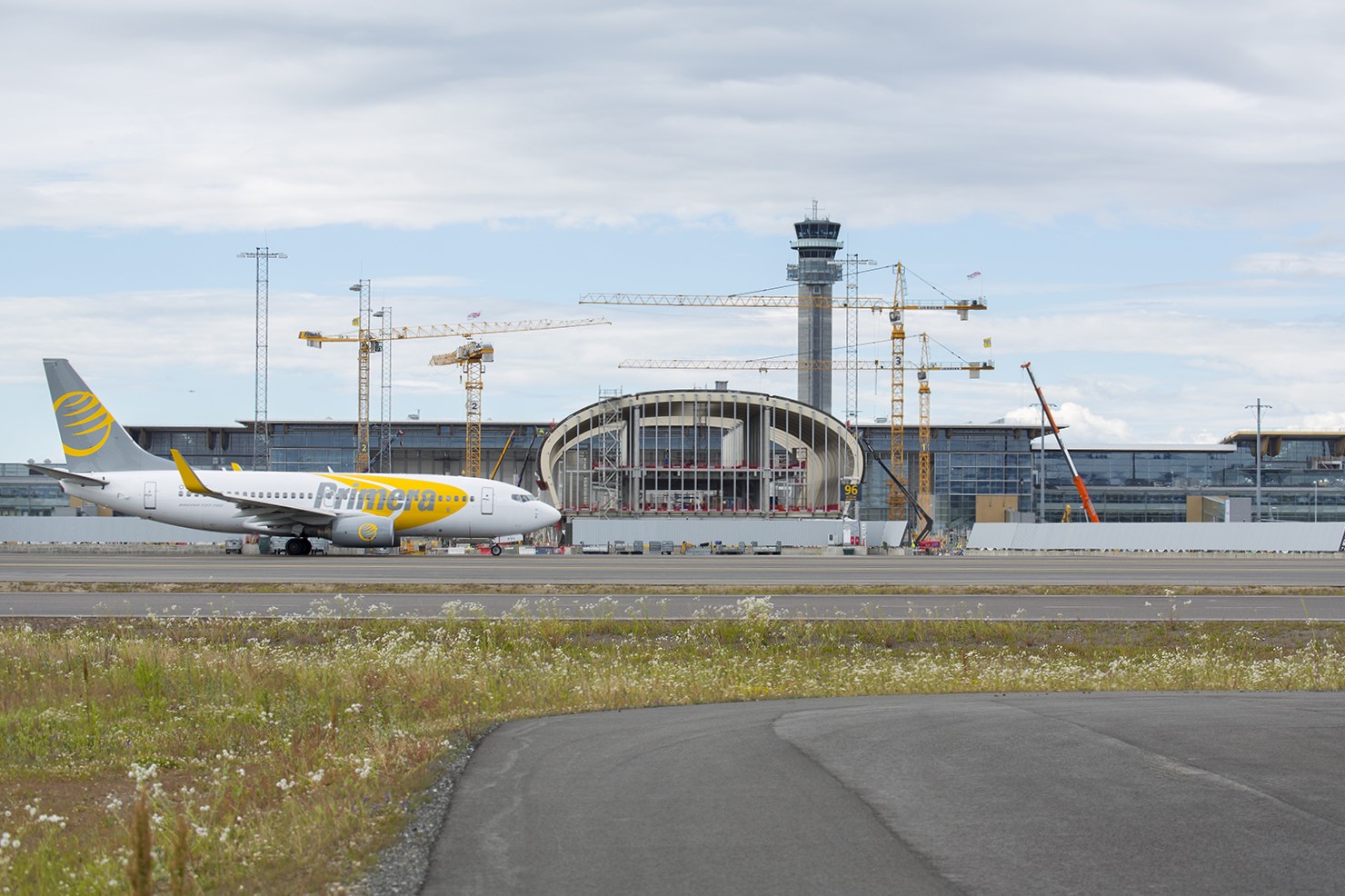 Flyplass med eit fly på rullebanen og byggearbeid i bakgrunnen. Kontrolltårn og fleire byggekranar er synlege under skyet himmel.