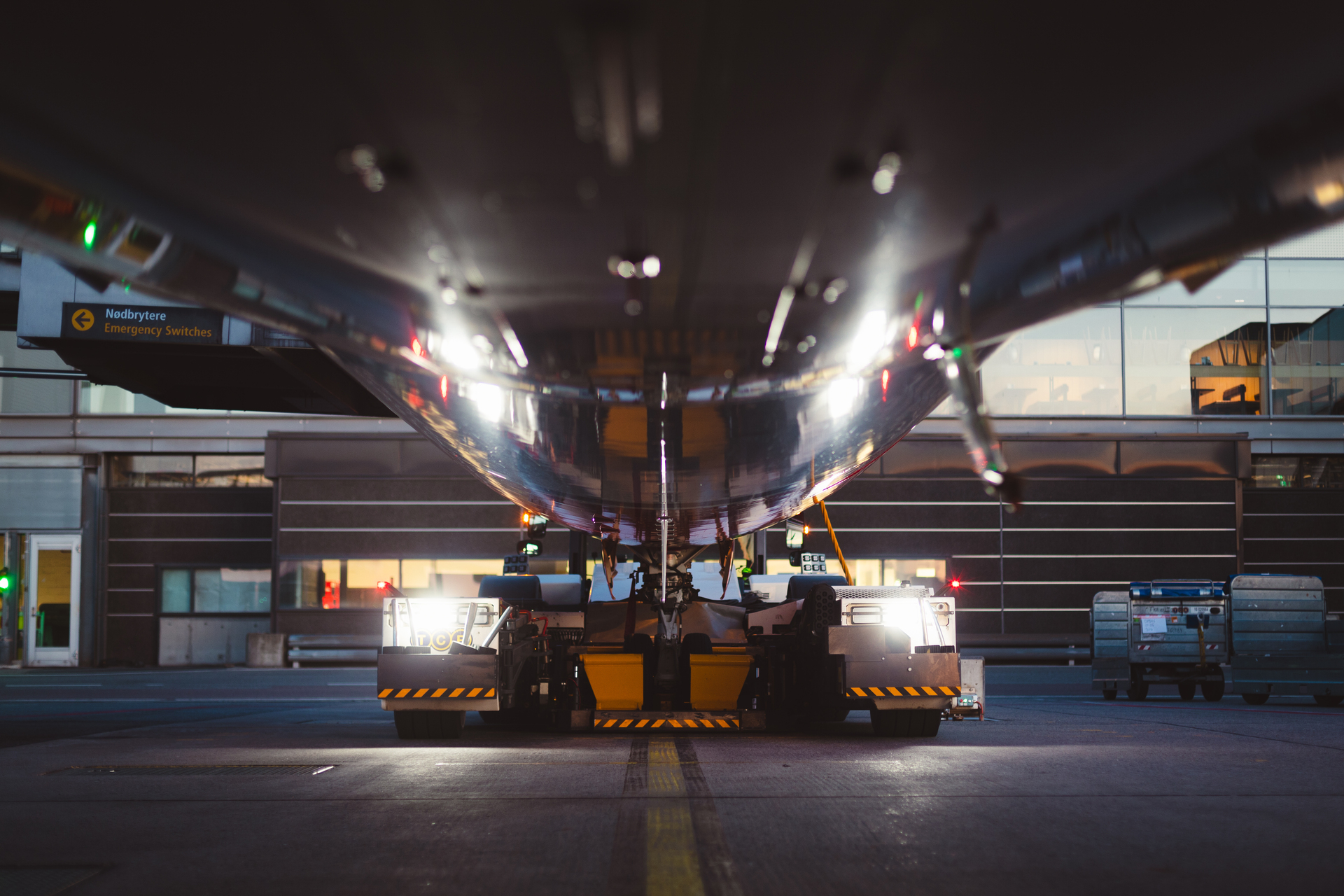 Underfra utsikt av et flyplassbakkekjøretøy som trekker et fly i skumringen, med refleksjoner på flyets overflate og terminalbygninger i bakgrunnen