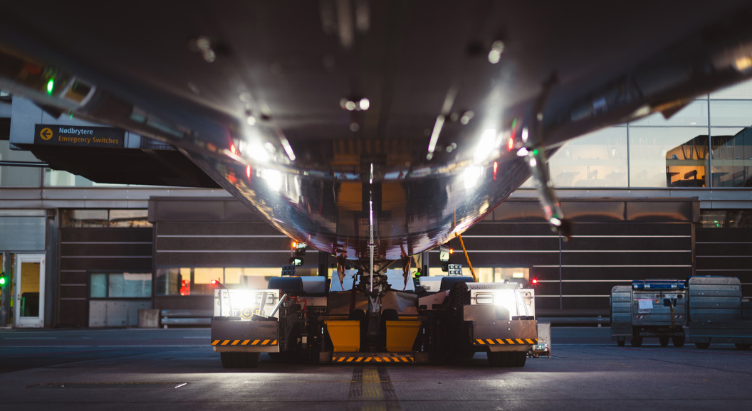 Underfra utsikt av et flyplassbakkekjøretøy som trekker et fly i skumringen, med refleksjoner på flyets overflate og terminalbygninger i bakgrunnen