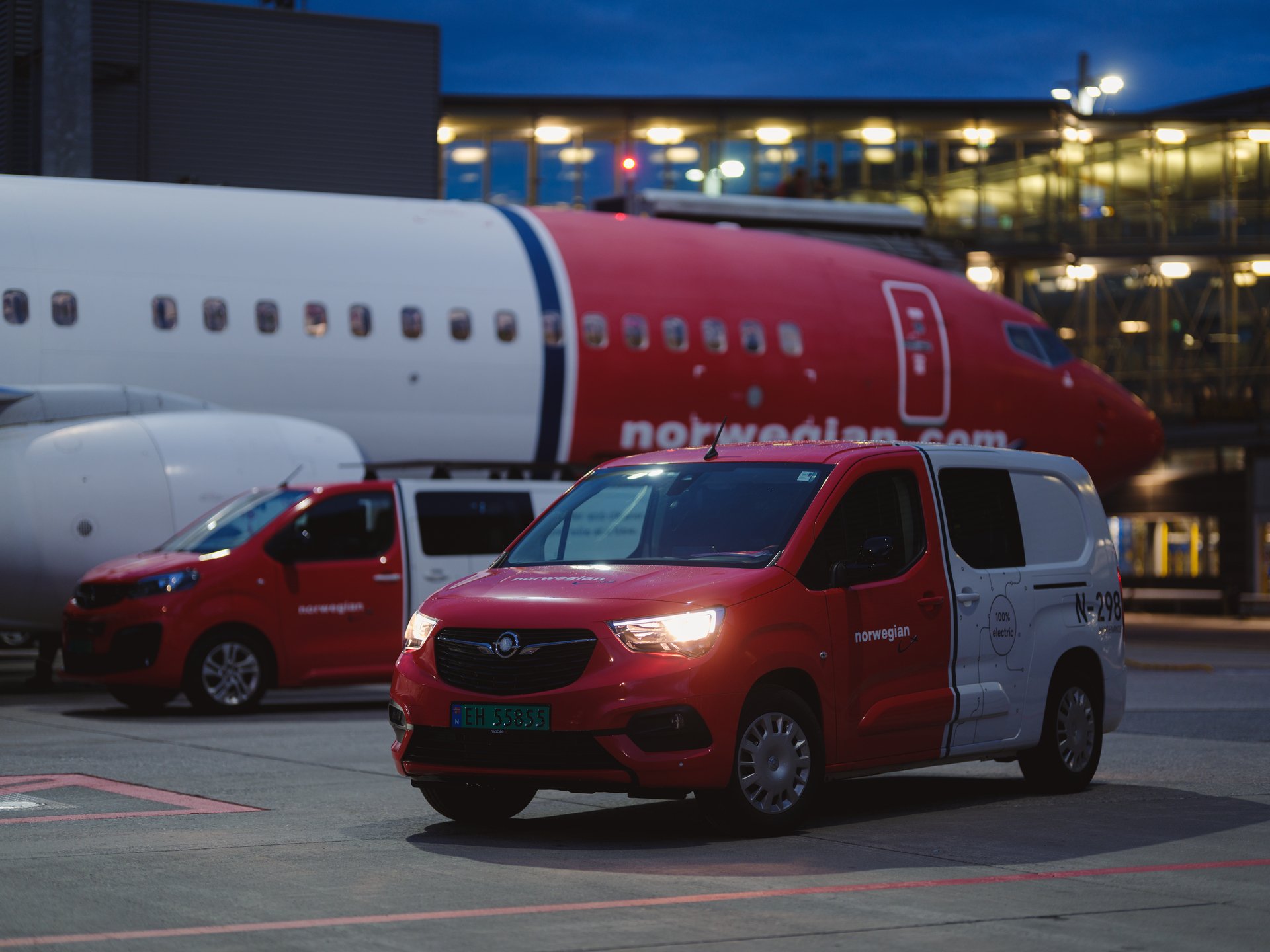 Røde og hvite servicebiler parkert nær et Norwegian Air Shuttle fly ved en flyplassterminal i kveldstimene