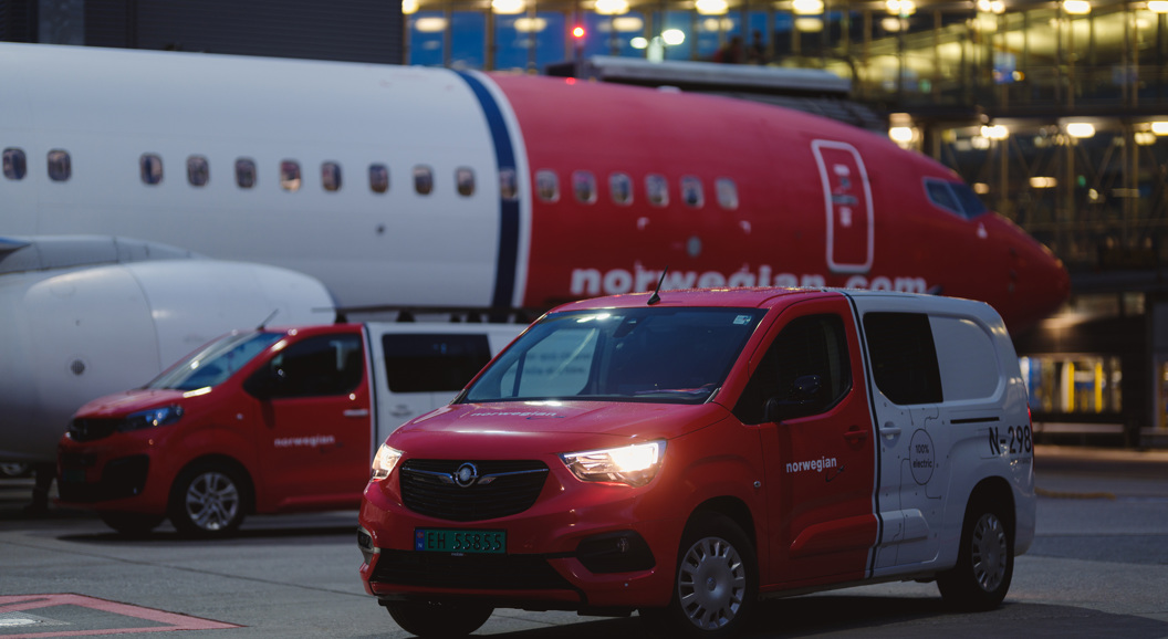 Røde og hvite servicebiler parkert nær et Norwegian Air Shuttle fly ved en flyplassterminal i kveldstimene