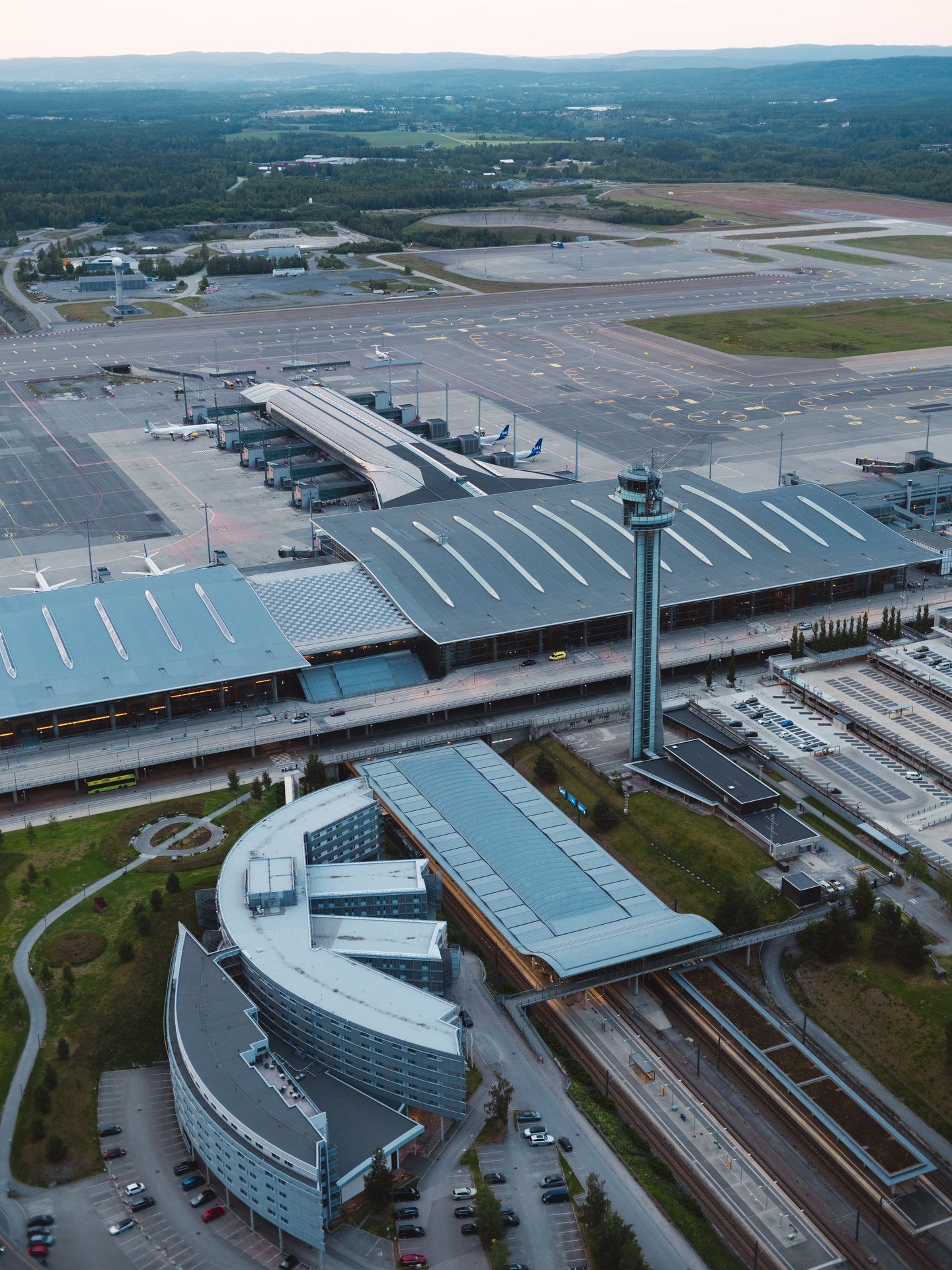 Flyplass med et stort passasjerfly parkert ved terminalen, byggkran i forgrunnen, og søknadsskjema med penn på bordet, symboliserer muligheter innen luftfartsindustrien.